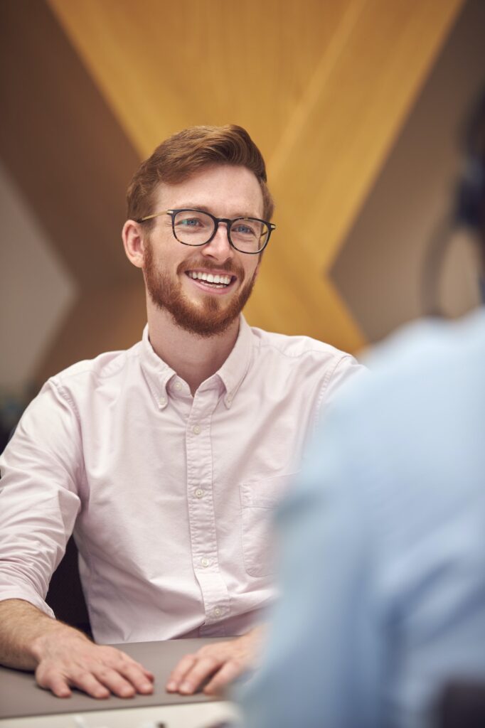 Businessman In Office Talking To Male Colleague Sitting At Desk